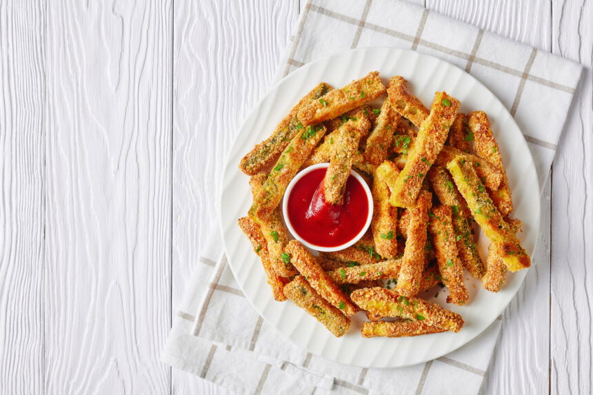 crunchy zucchini sticks breaded with panko breadcrumbs, parmesan cheese, spices on a white plate with ketchup on a wooden table